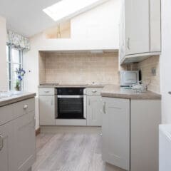 Bright, modern kitchen with light-colored cabinets, wood-style flooring, beige tile backsplash, built-in oven, microwave, sink by the window, and natural light from a skylight above—ideal for home refurbishments or loft conversions.