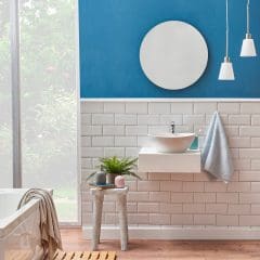 A modern bathroom by an expert builder features blue and white walls, a round mirror, white sink with towel, pendant lights, a wooden stool with plants and candles, a bathtub, and a wooden bath mat on light wood flooring.