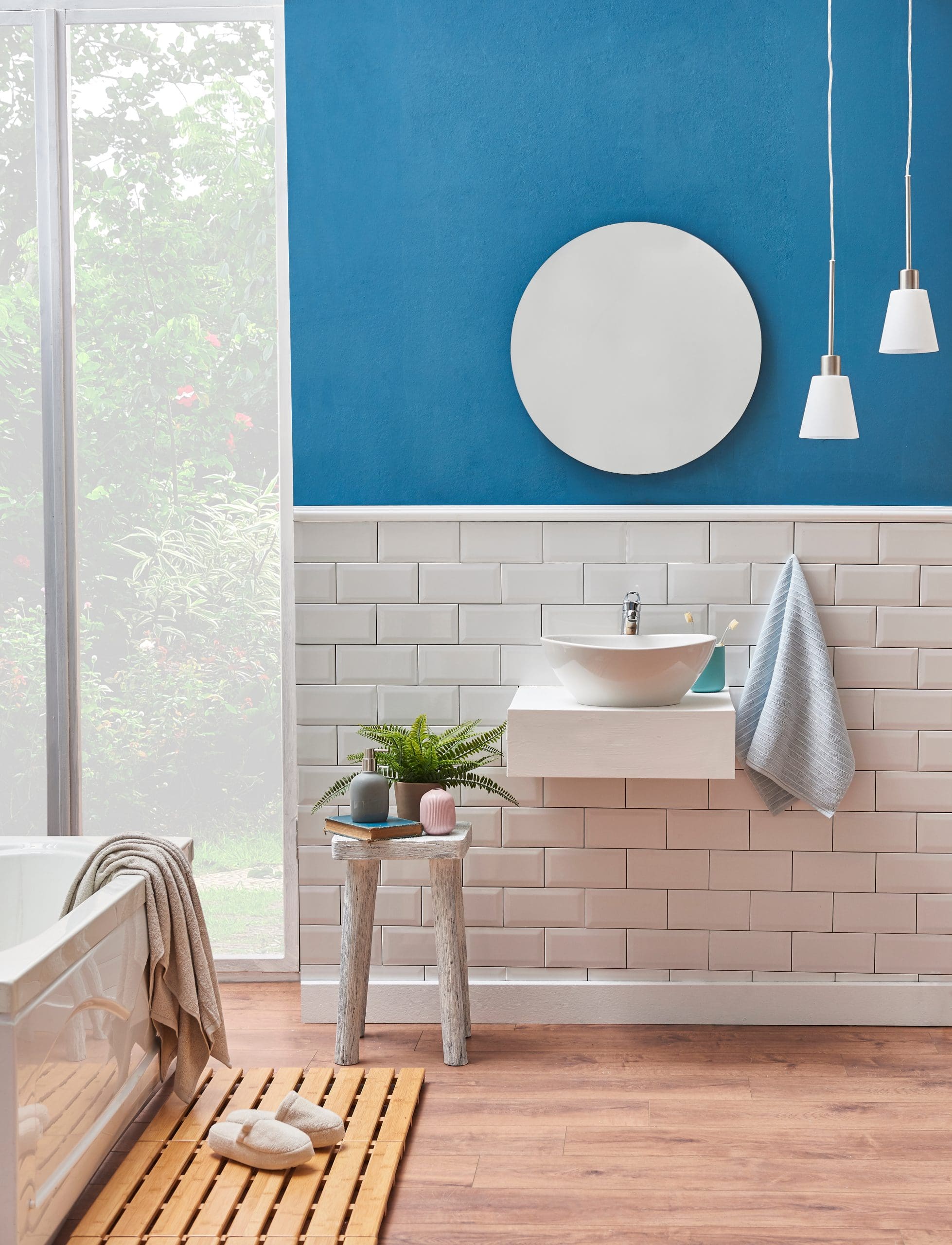 A modern bathroom by an expert builder features blue and white walls, a round mirror, white sink with towel, pendant lights, a wooden stool with plants and candles, a bathtub, and a wooden bath mat on light wood flooring.
