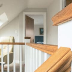 Close-up of a wooden handrail on a staircase landing in a bright, modern home with white walls and a slanted ceiling—showcasing the quality craftsmanship possible with house extensions and home refurbishments. An open door and beige armchair appear in the background.