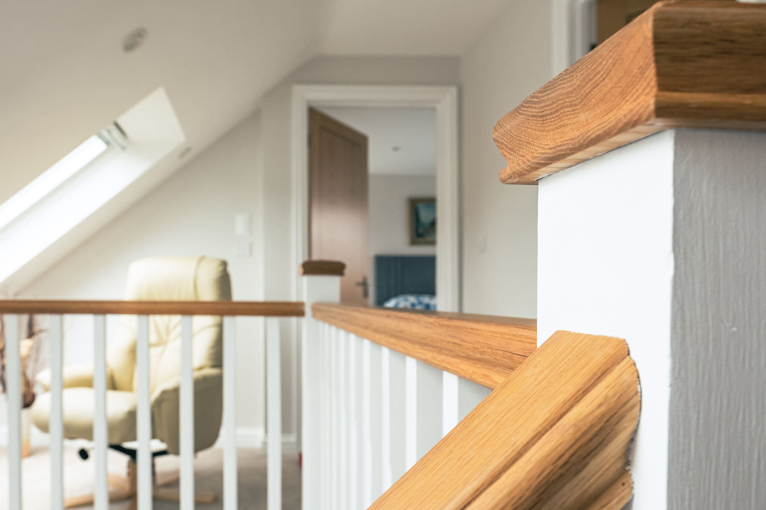 Close-up of a wooden handrail on a staircase landing in a bright, modern home with white walls and a slanted ceiling—showcasing the quality craftsmanship possible with house extensions and home refurbishments. An open door and beige armchair appear in the background.