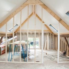 A partially finished attic space with exposed wooden framing, construction materials, tools, and insulation scattered around—ideal for Loft Conversions. Sunlight enters through skylight windows on the sloped ceiling.