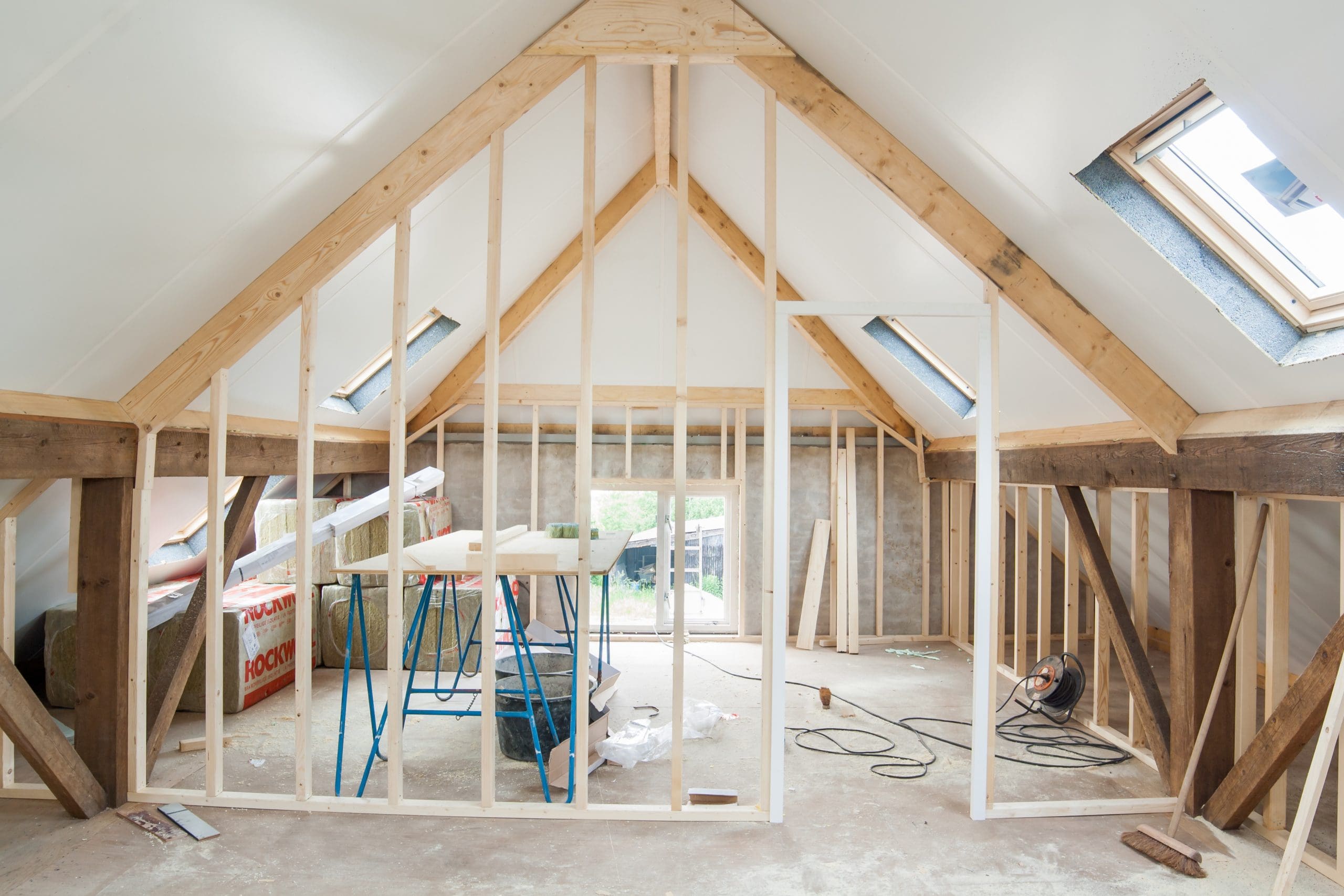 A partially finished attic space with exposed wooden framing, construction materials, tools, and insulation scattered around—ideal for Loft Conversions. Sunlight enters through skylight windows on the sloped ceiling.