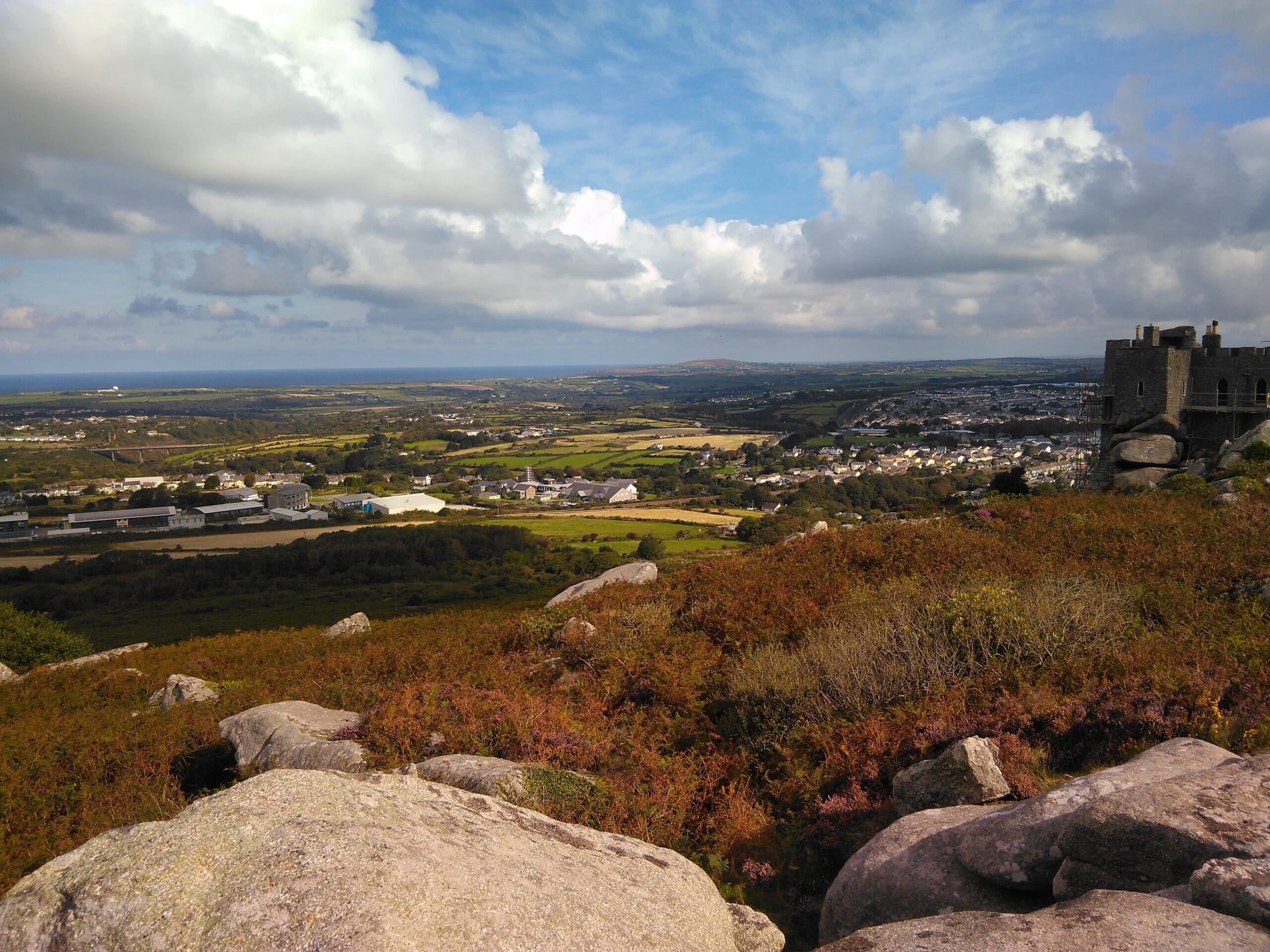 A scenic view from a rocky hilltop shows a patchwork of green fields, small buildings, and a partially visible stone structure—perhaps awaiting home refurbishments by a skilled builder—under a partly cloudy sky.