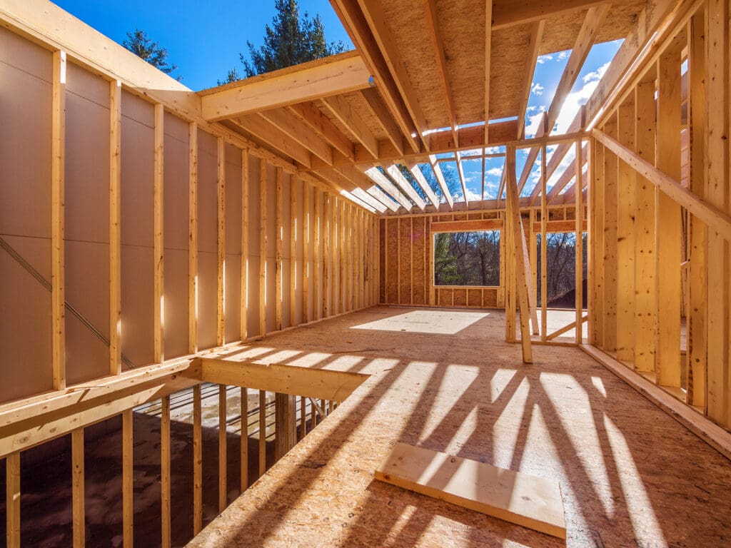 Sunlit interior of a wooden house under construction showcases exposed framing and open ceiling beams—an inspiring scene for new house building or future loft conversions, with blue sky and trees visible through the openings.