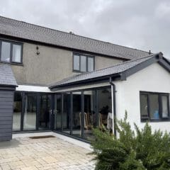 A modern two-story house with gray roof tiles, light gray stucco walls, large glass sliding doors, and a paved patio—perfect inspiration for new house building. Greenery fills the foreground beneath a cloudy sky.